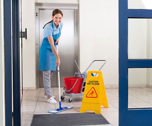 A cleaner mopping a communal hallway beside the lift, with a mop bucket and wet floor caution sign
